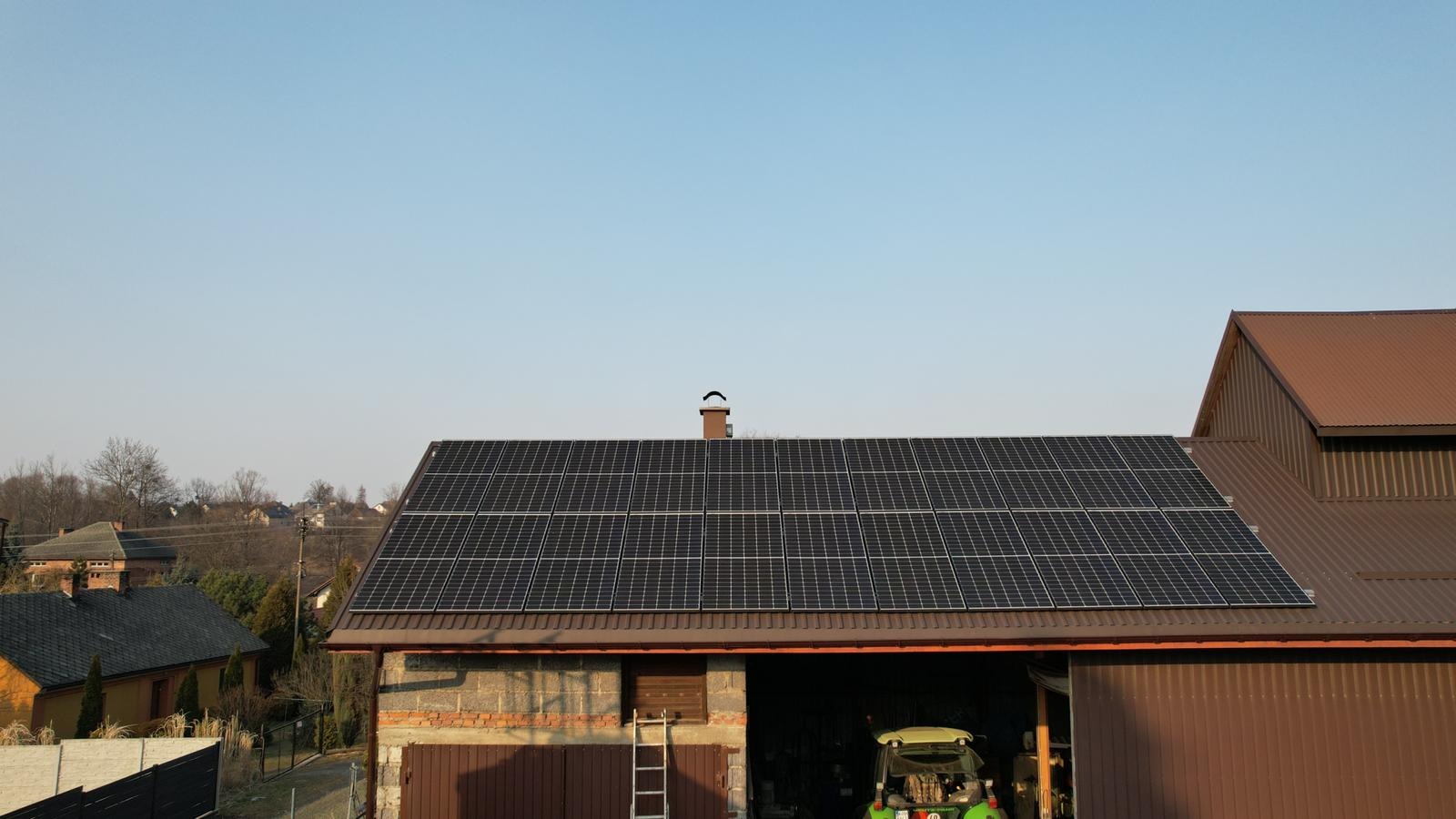 Panele fotowoltaiczne na dachu budynku gospodarczego z cegły, z traktorem w garażu i drabiną opartą o ścianę.