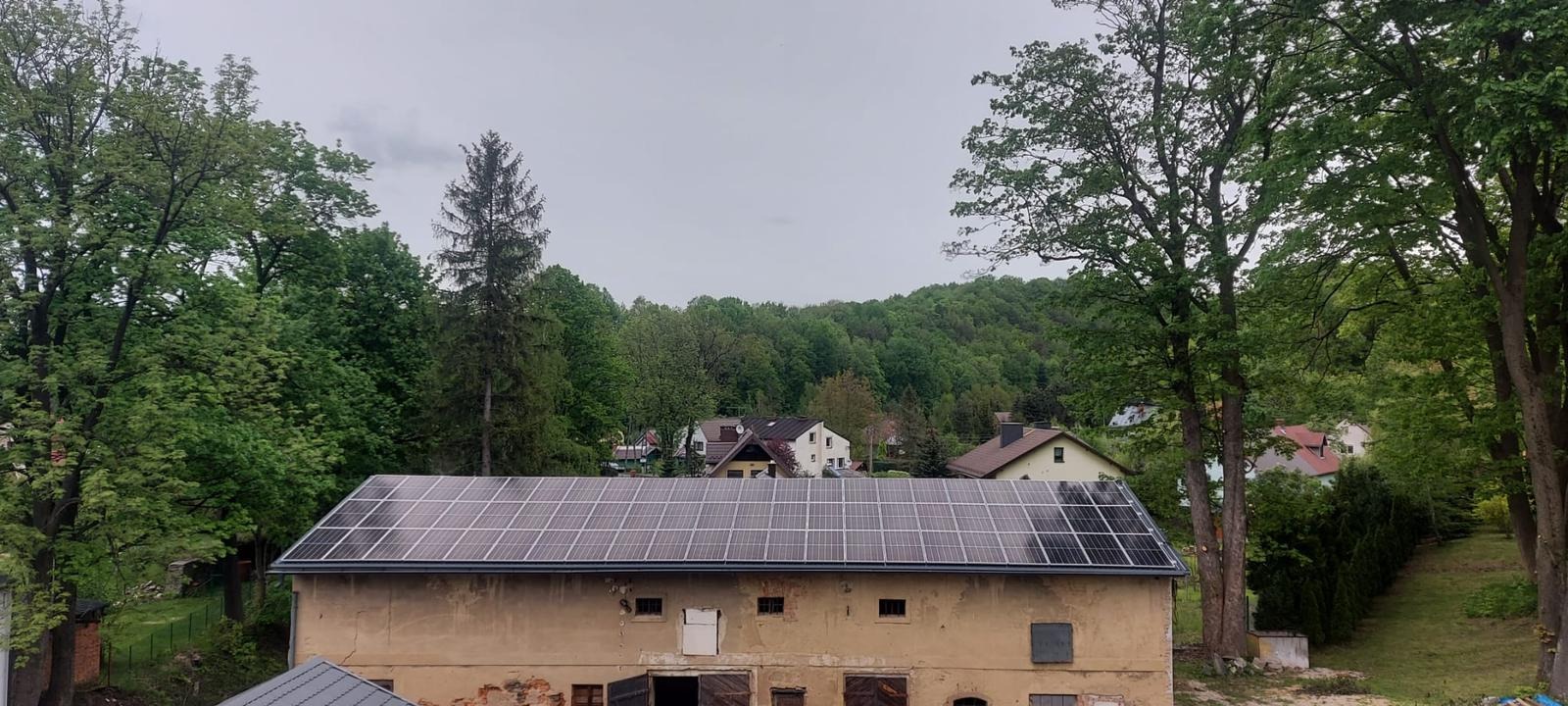 Panele słoneczne na dachu starego budynku gospodarczego, widok z góry na tle zieleni i zabudowań wiejskich w pochmurny dzień.