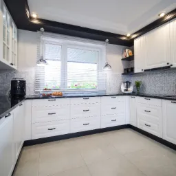 Jasna kuchnia w stylu klasycznym z białymi szafkami, czarnymi blatami i geometryczną mozaiką na ścianie, doświetlona lampami wiszącymi i światłem naturalnym z okna z widokiem na zieleń.