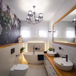 Nowoczesna łazienka z wolnostojącą wanną, dwiema owalnymi umywalkami na drewnianym blacie, tapetą w kwiaty i designerskim żyrandolem