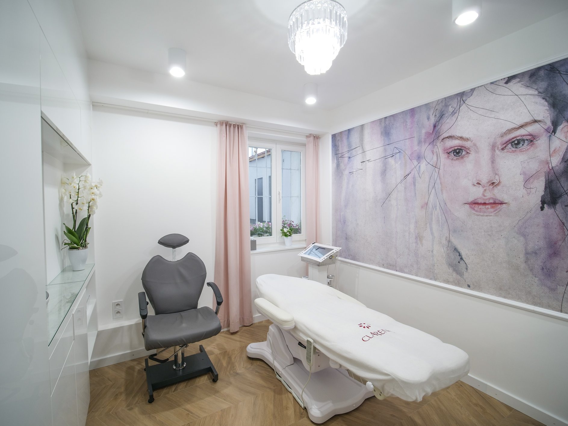 Jasne wnętrze gabinetu kosmetycznego z regulowanym fotelem, łóżkiem zabiegowym z logo Clarena, akwarelą portretową na ścianie i kwiatami na parapecie.
