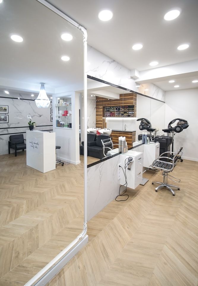 Jasne wnętrze salonu fryzjerskiego z recepcją, stanowiskami do mycia włosów i suszarkami hełmowymi, wykończone marmurem i drewnem, widziane przez lustro z ozdobną ramą.