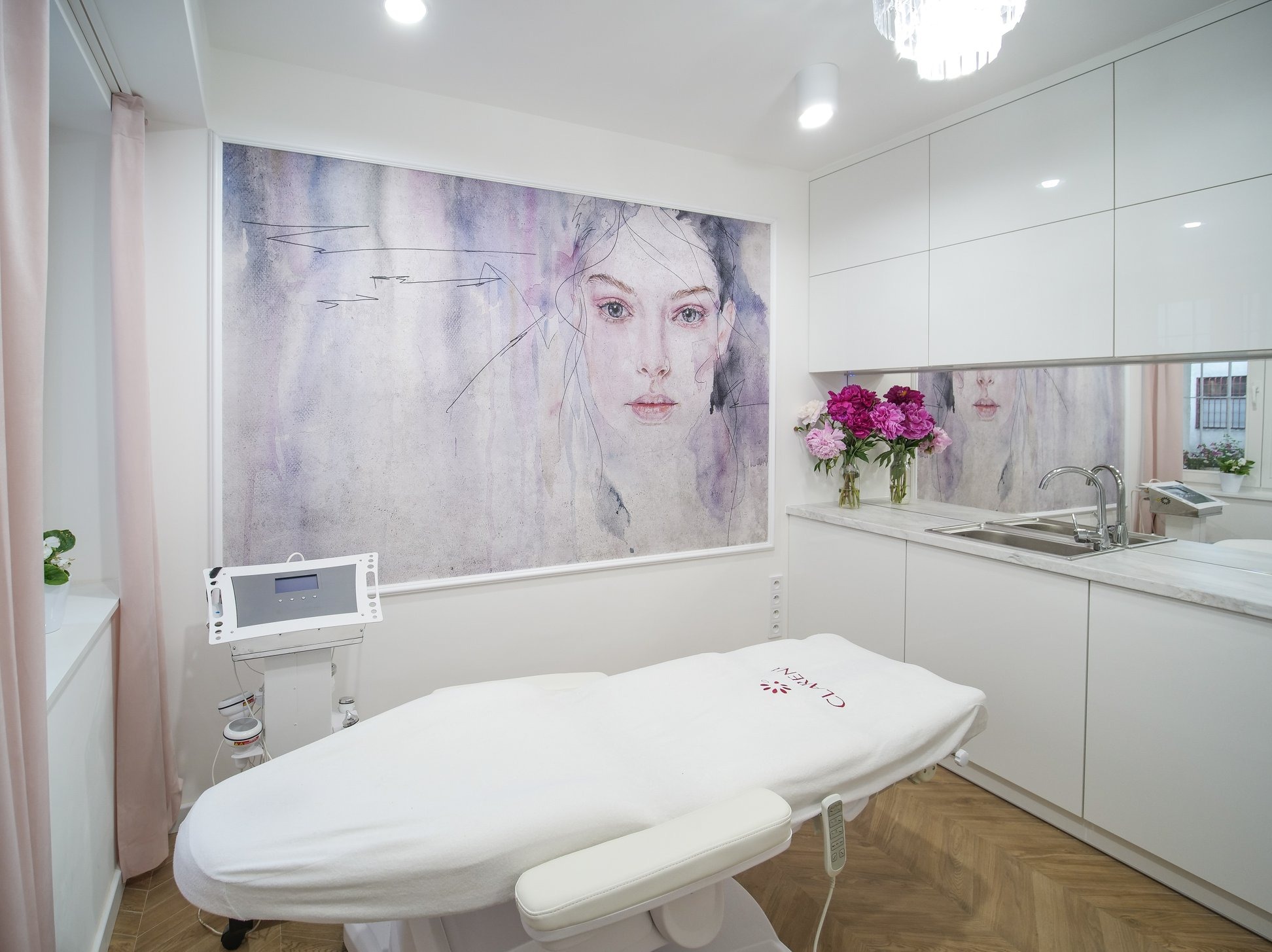 Jasne wnętrze gabinetu kosmetycznego z łóżkiem do zabiegów, aparaturą kosmetyczną, grafiką na ścianie i bukietem piwonii.
