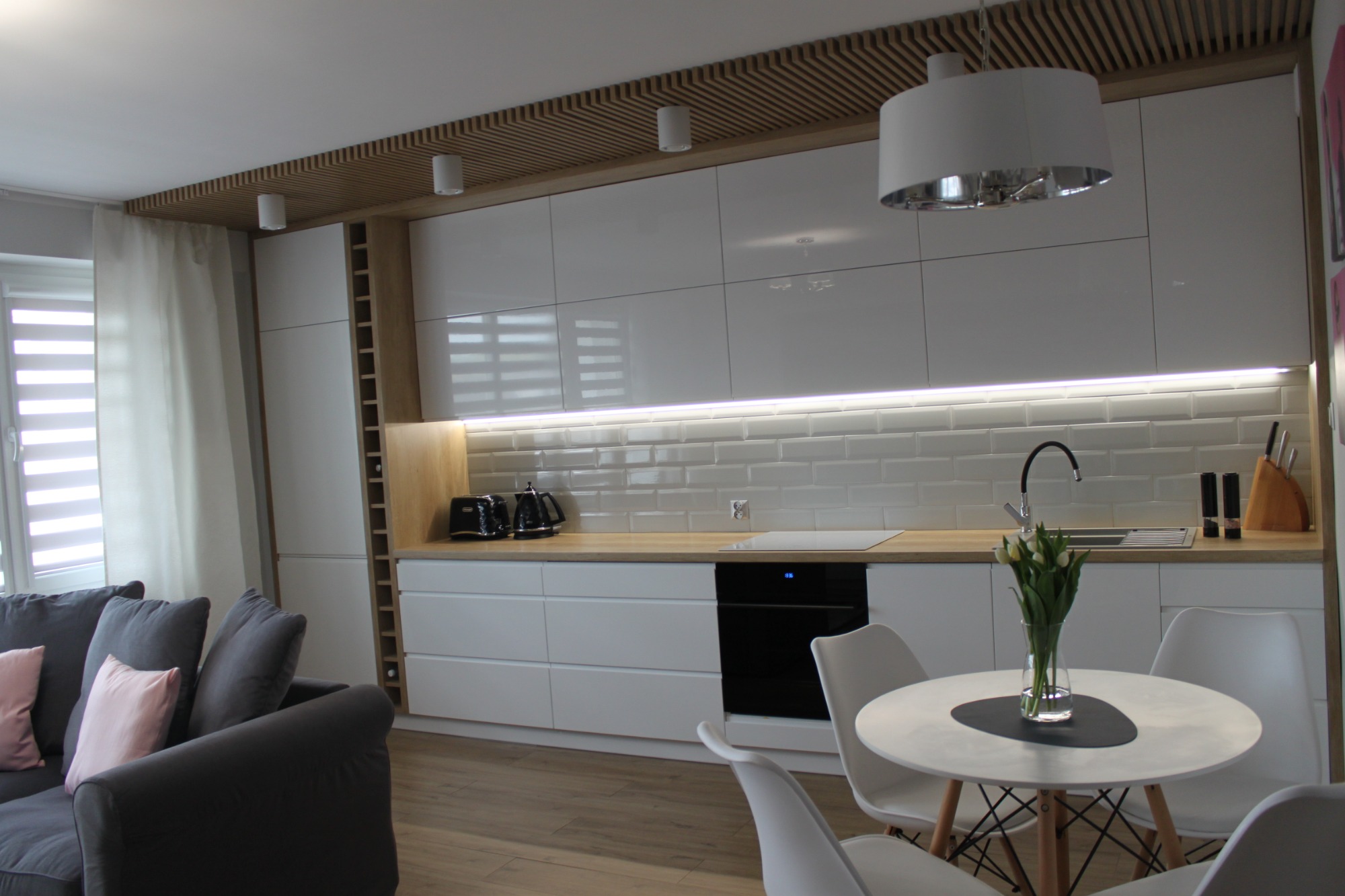 Nowoczesna kuchnia z białymi, błyszczącymi szafkami, drewnianym blatem i elementami dekoracyjnymi z drewna na suficie, uzupełniona o szary narożnik z różową poduszką w tle.