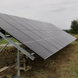 Panel fotowoltaiczny zamontowany na metalowej konstrukcji w otoczeniu trawy i drzew, widoczne ślady prac ziemnych.