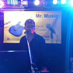 Mr. Music - Wokalista zespołu muzycznego Mr. Music przed banerem reklamowym z klawiaturą i gitarą elektryczną, oświetlony kolorowymi światłami podczas występu.