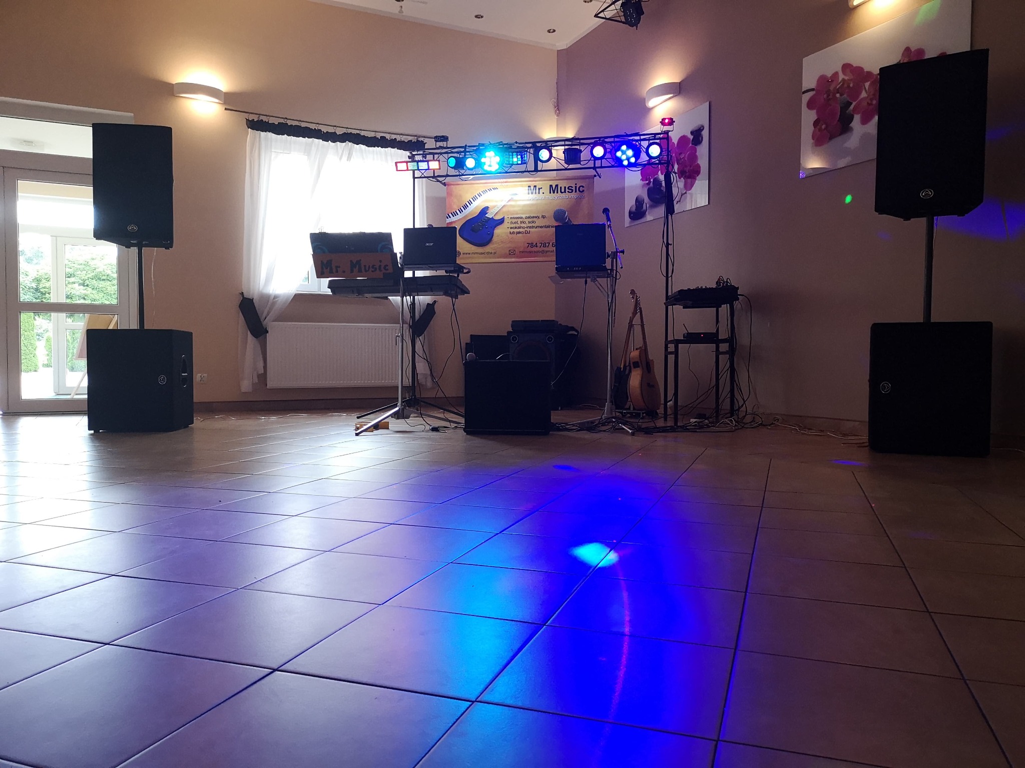 Scena z nagłośnieniem i oświetleniem przygotowana do występu, widoczny baner reklamowy z nazwą 'Mr. Music' i informacjami kontaktowymi, gitara oparta o statyw. Podłoga oświetlona niebieskim światłem.