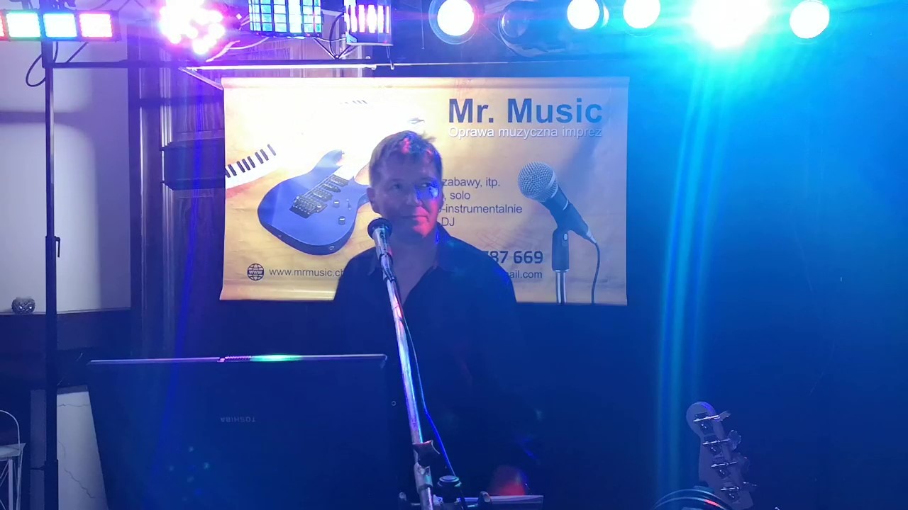 Wokalista zespołu muzycznego Mr. Music przed banerem reklamowym z klawiaturą i gitarą elektryczną, oświetlony kolorowymi światłami podczas występu.