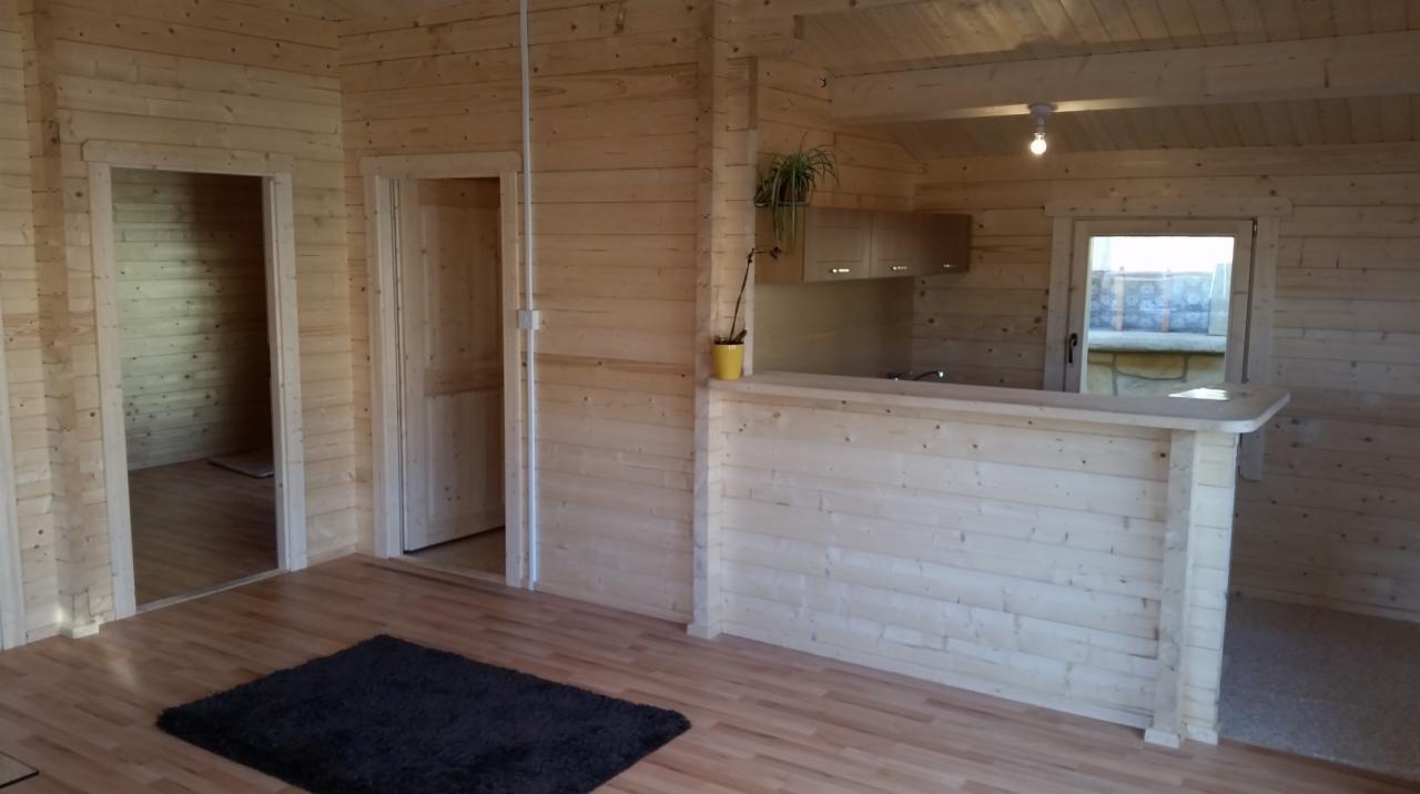 Wnętrze domu z drewnianych bali z widoczną otwartą kuchnią z barem, drewnianą podłogą i czarnym dywanem, z dwoma otworami drzwiowymi prowadzącymi do innych pomieszczeń.