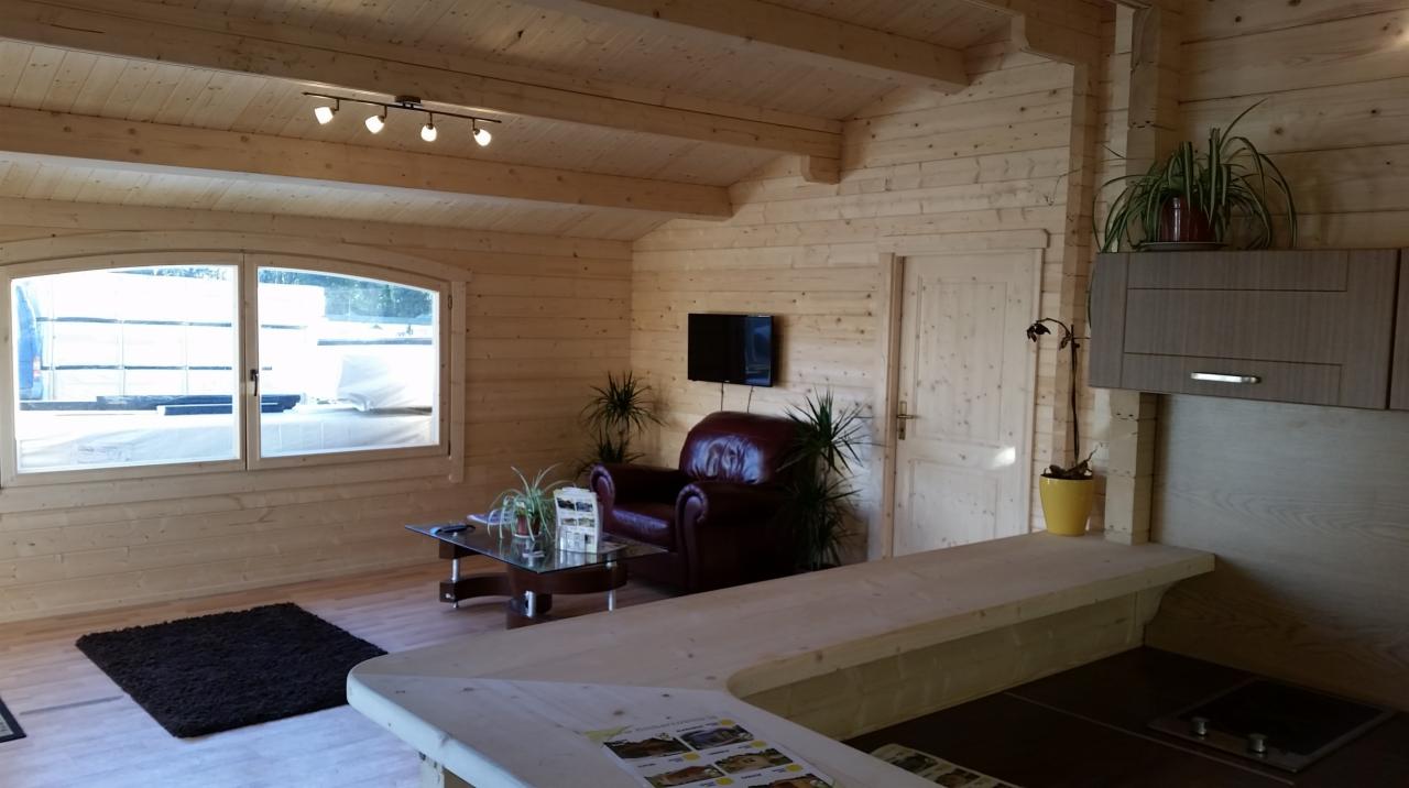 Wnętrze drewnianego domu z bali w Warszawie, z widocznym salonem z brązowym fotelem i aneksem kuchennym, wykończone jasnym drewnem.