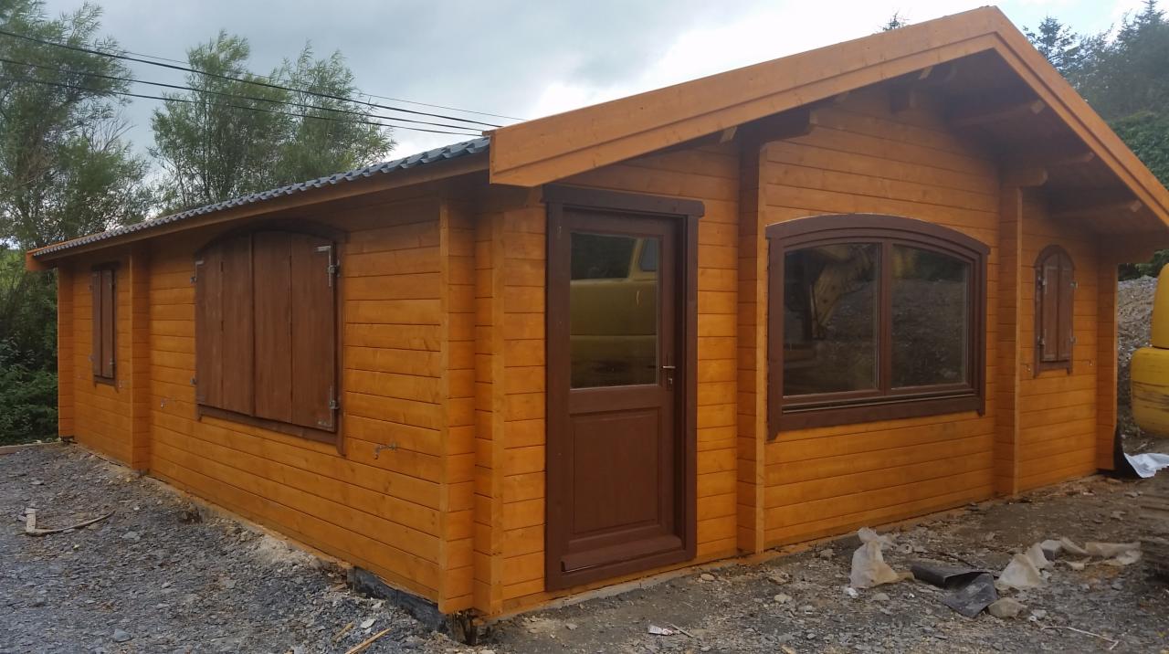 Gotowy, parterowy budynek z bali z ciemnobrązowymi oknami i drzwiami, widoczne ślady budowy na ziemi.