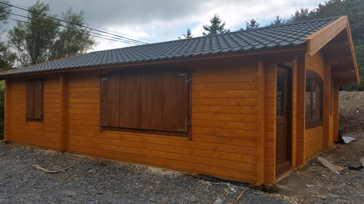 Zewnętrzny widok świeżo postawionego, parterowego domu z bali z ciemnymi, zamkniętymi okiennicami, ciemną dachówką i widocznymi śladami prac budowlanych wokół.