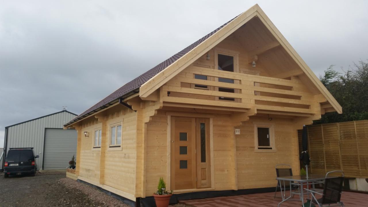 Nowoczesny, parterowy dom z jasnego drewna z balkonem i brązowym dachem, na zewnątrz widoczny stolik z krzesłami i mała choinka w doniczce.