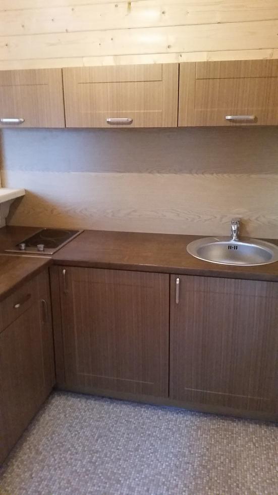 Wnętrze kuchni w domu z bali, z drewnianymi szafkami, płytą indukcyjną i okrągłym zlewozmywakiem ze stali nierdzewnej, wykończone drewnem na ścianach i mozaikową podłogą.