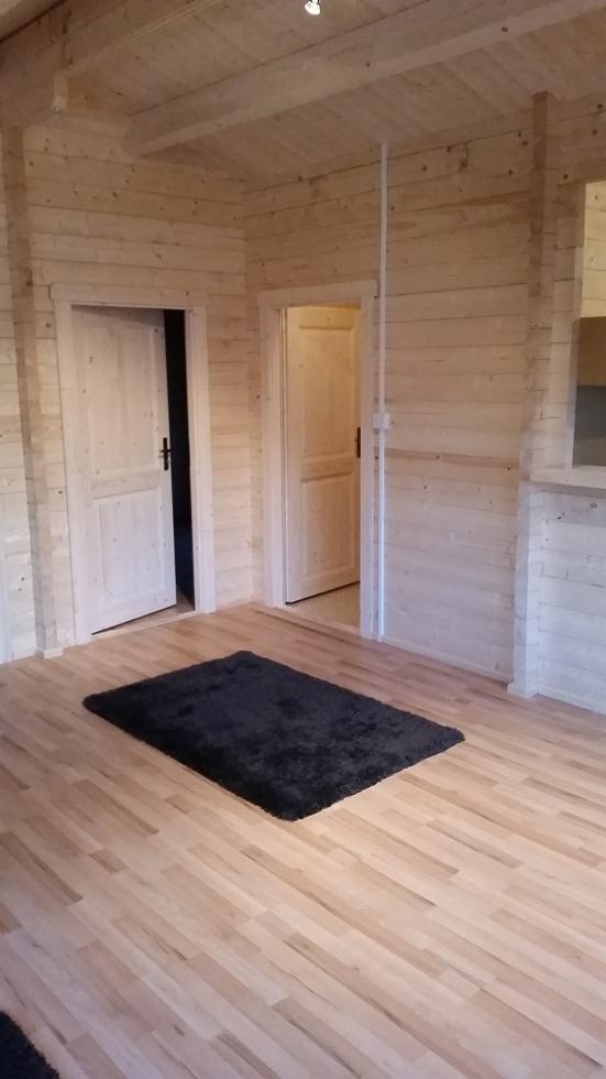 Wnętrze pokoju wykończonego jasnym drewnem, z widocznymi dwoma białymi drzwiami z czarnymi klamkami oraz ciemnym, prostokątnym dywanem na podłodze.