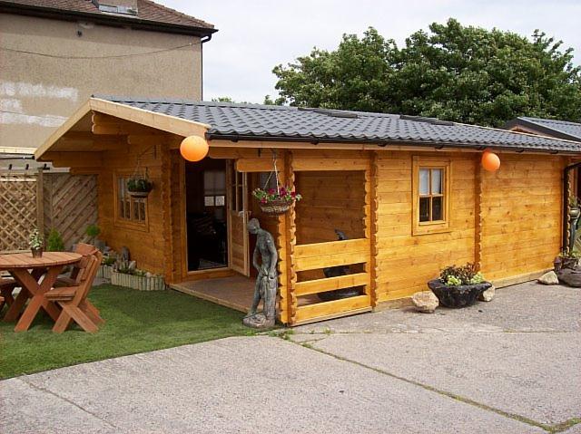 Jasny, parterowy budynek z bali z szarą dachówką, ozdobiony wiszącymi kwiatami i pomarańczowymi kulami, z drewnianym stołem i krzesłami na trawie oraz rzeźbą przy wejściu.