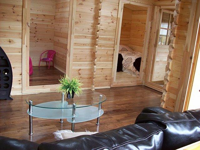 Wnętrze domu z bali z drewnianymi ścianami, widoczne dwa małe pokoje z wyposażeniem, szklany stolik kawowy z rośliną doniczkową i fragment czarnej skórzanej kanapy w salonie.