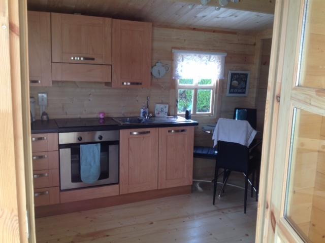 Wnętrze kuchni w drewnianym domku, z jasnymi szafkami, oknem z firanką i widokiem na zieleń, z ręcznikiem na piekarniku i krzesłem z białym materiałem w tle.