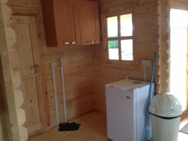 Wnętrze drewnianego domku: widoczne szafki wiszące, okno z widokiem na zewnątrz, mała lodówka, kosz na śmieci i instalacja elektryczna na ścianie.