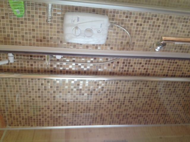 Niewyraźne ujęcie kabiny prysznicowej z mozaiką w odcieniach brązu, widoczny biały podgrzewacz wody i chromowana armatura.