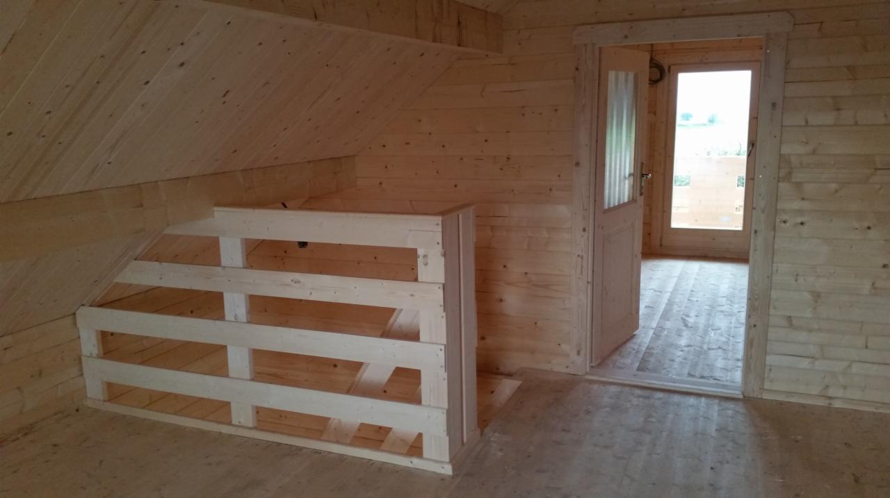 Wnętrze surowego, drewnianego domu z bali w trakcie budowy, widoczne drewniane schody bez poręczy prowadzące na antresolę i otwarte drzwi z widokiem na zewnątrz.