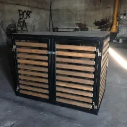 Metalowa szafka z drewnianymi lamelami na drzwiach, zamykana na zasuwkę, w surowym, warsztatowym otoczeniu z betonową podłogą i ścianami z graffiti.