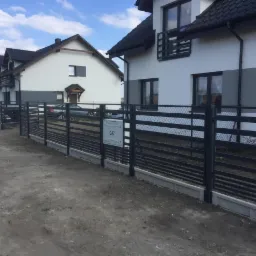 Nowoczesne, poziome przęsła ogrodzeniowe w kolorze grafitowym, z siatką na górze, zamontowane na betonowej podmurówce, przed dwoma nowymi domami z szarobiałą elewacją i czarnymi dachami.