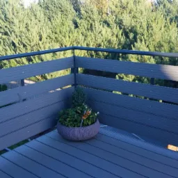 Szara balustrada balkonowa z kompozytu drewna, czarne metalowe słupki, donica z kwiatami na tle iglastego lasu.