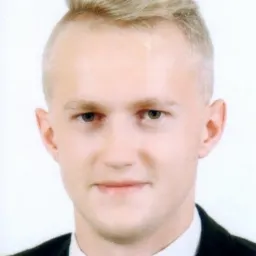 Portret młodego mężczyzny w garniturze, z jasnymi włosami zaczesanymi do góry i lekko uśmiechniętą twarzą na białym tle.