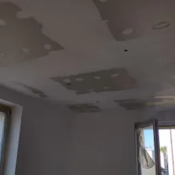 Nierówna powierzchnia sufitu z widocznymi łatami po szpachlowaniu, przygotowana do dalszych prac wykończeniowych w jasnym pomieszczeniu z otwartym oknem. Fragment widoku na ulicę.