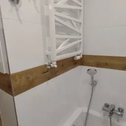 Biała wanna z chromowaną armaturą, panel dekoracyjny imitujący drewno, grzejnik drabinkowy w nowoczesnej łazience wykończonej białymi płytkami.