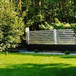 Nowoczesne ogrodzenie poziome z białymi sztachetami na ciemnej podmurówce z cegły, otoczone zielonym trawnikiem, drzewami i elementami architektury ogrodowej, takimi jak altana i ozdobne krzewy.