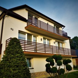 Hot Spaw SM Sebastian Moździerz - Dwupiętrowy dom z beżową elewacją, brązowymi balustradami balkonowymi i numerem '62' na ścianie, otoczony zielenią.