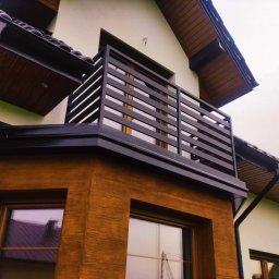 Hot Spaw SM Sebastian Moździerz - Czarna, pozioma balustrada balkonowa z metalowymi szczeblami, widok z dołu na tle elewacji domu z oknami.