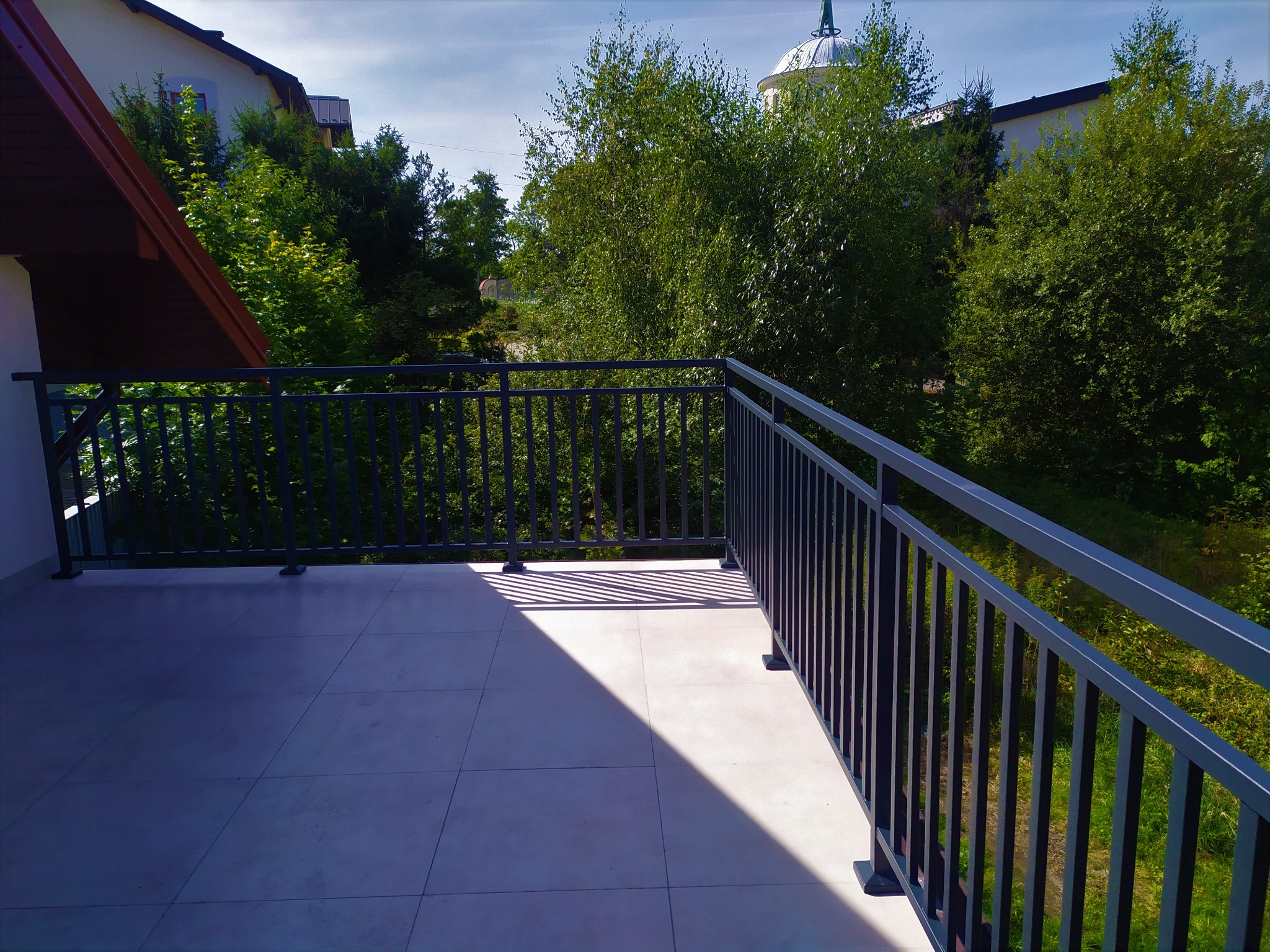 Nowoczesna, grafitowa balustrada balkonowa z pionowymi szczebelkami w Dąbrowie Tarnowskiej, z widokiem na zieleń i fragment zabudowań w tle.