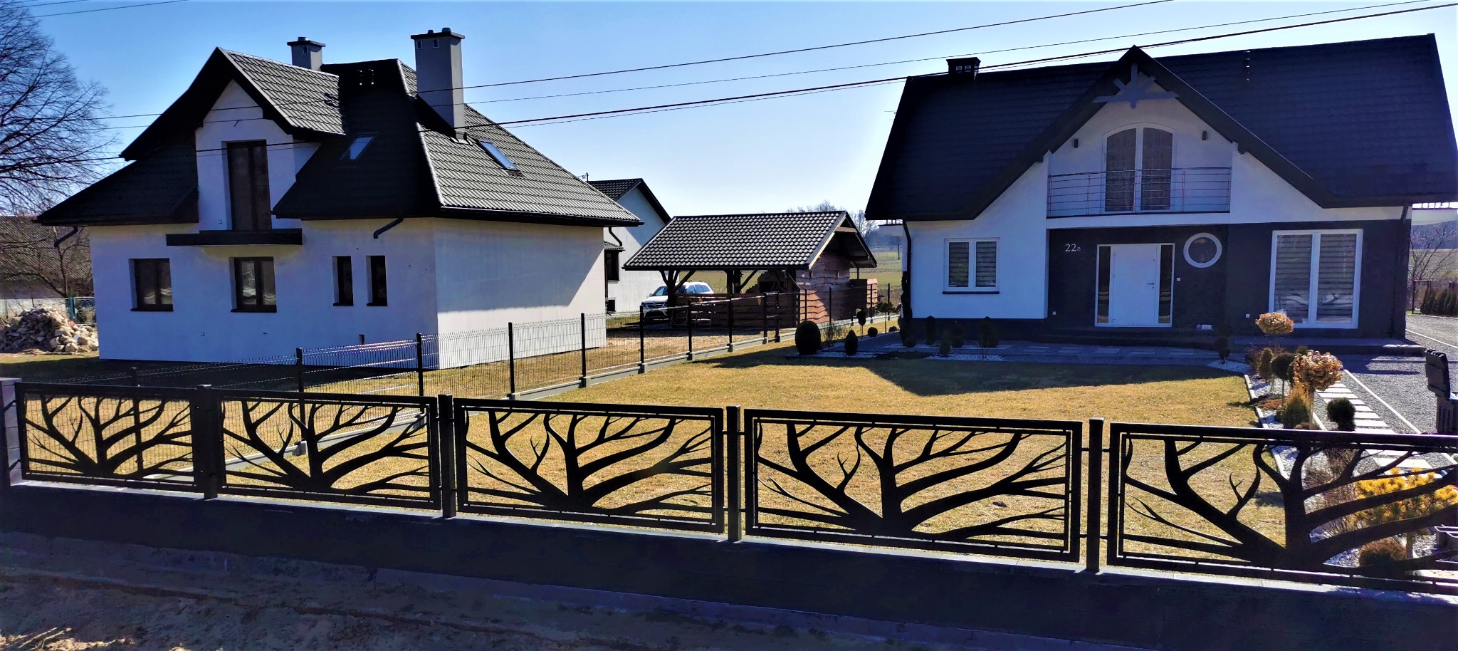 Nowoczesne ogrodzenie z metalu z motywem drzewa przed domem z białymi ścianami i ciemnym dachem w słoneczny dzień.