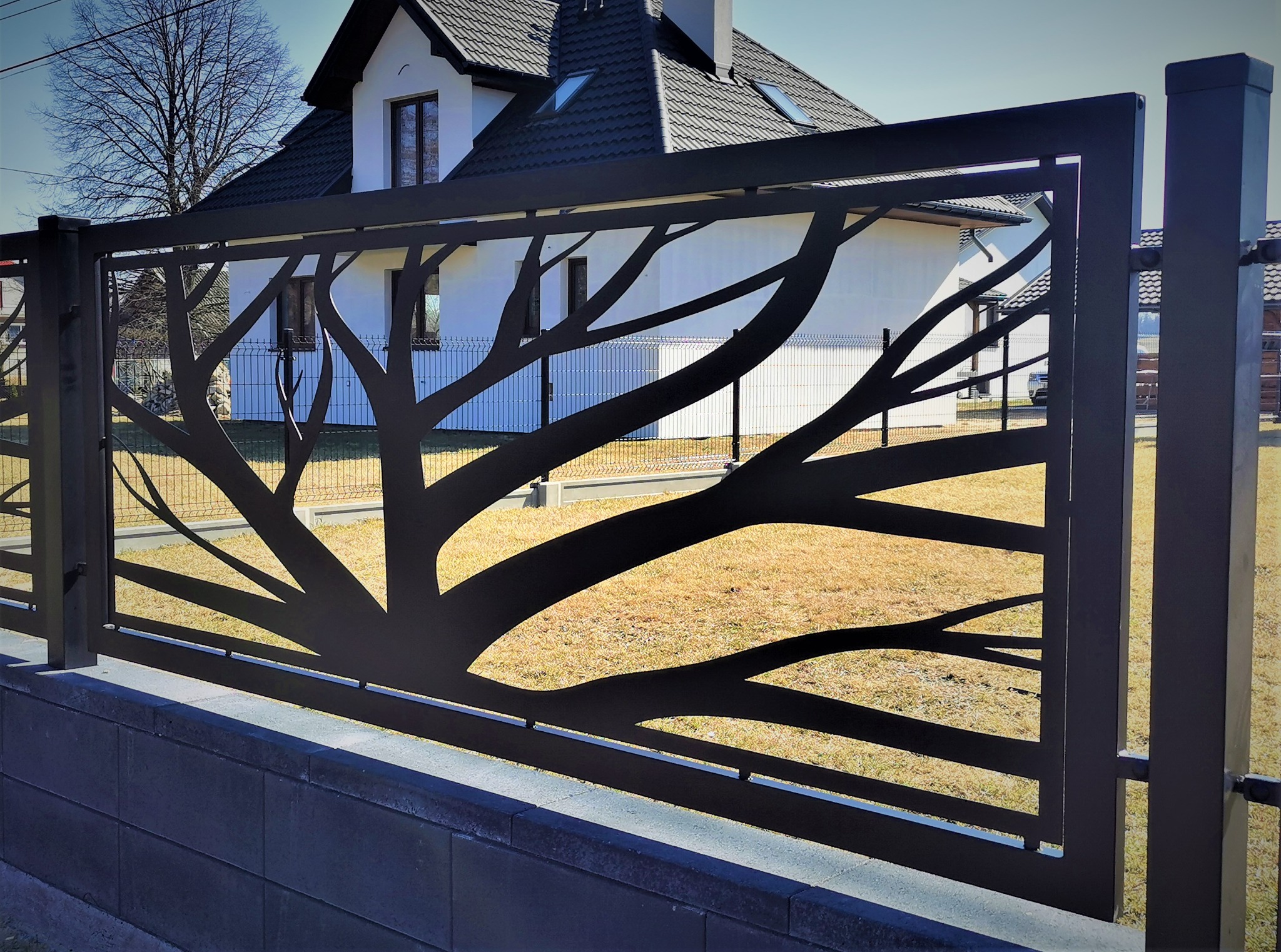 Nowoczesne ogrodzenie panelowe z motywem drzewa na tle domu z białymi ścianami i ciemną dachówką.