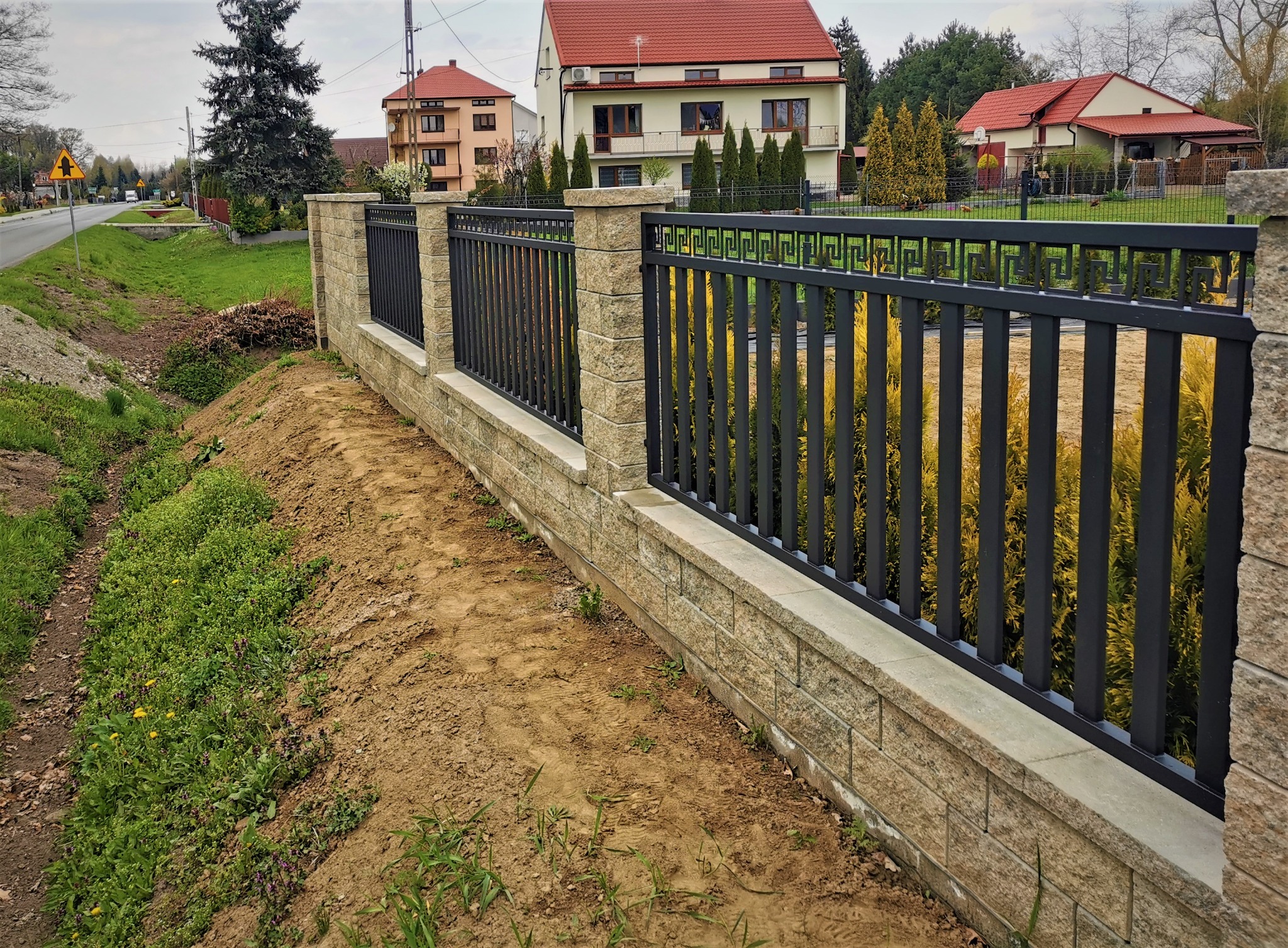 Nowoczesne ogrodzenie z czarnymi, pionowymi prętami i beżowymi słupkami z bloczków betonowych, oddzielające posesję od drogi z widocznymi domami w tle.