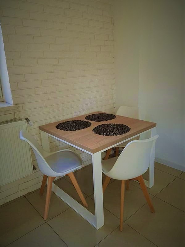 Stół z jasnego drewna z trzema czarnymi podkładkami i białymi krzesłami z drewnianymi nogami ustawiony w rogu pokoju z ceglaną ścianą.