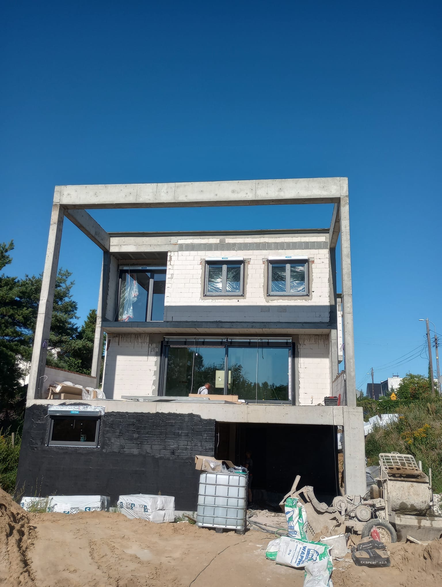 Surowy stan dwukondygnacyjnego domu z betonu komórkowego i ramą betonową w słoneczny dzień. Widoczne okna i materiały budowlane.