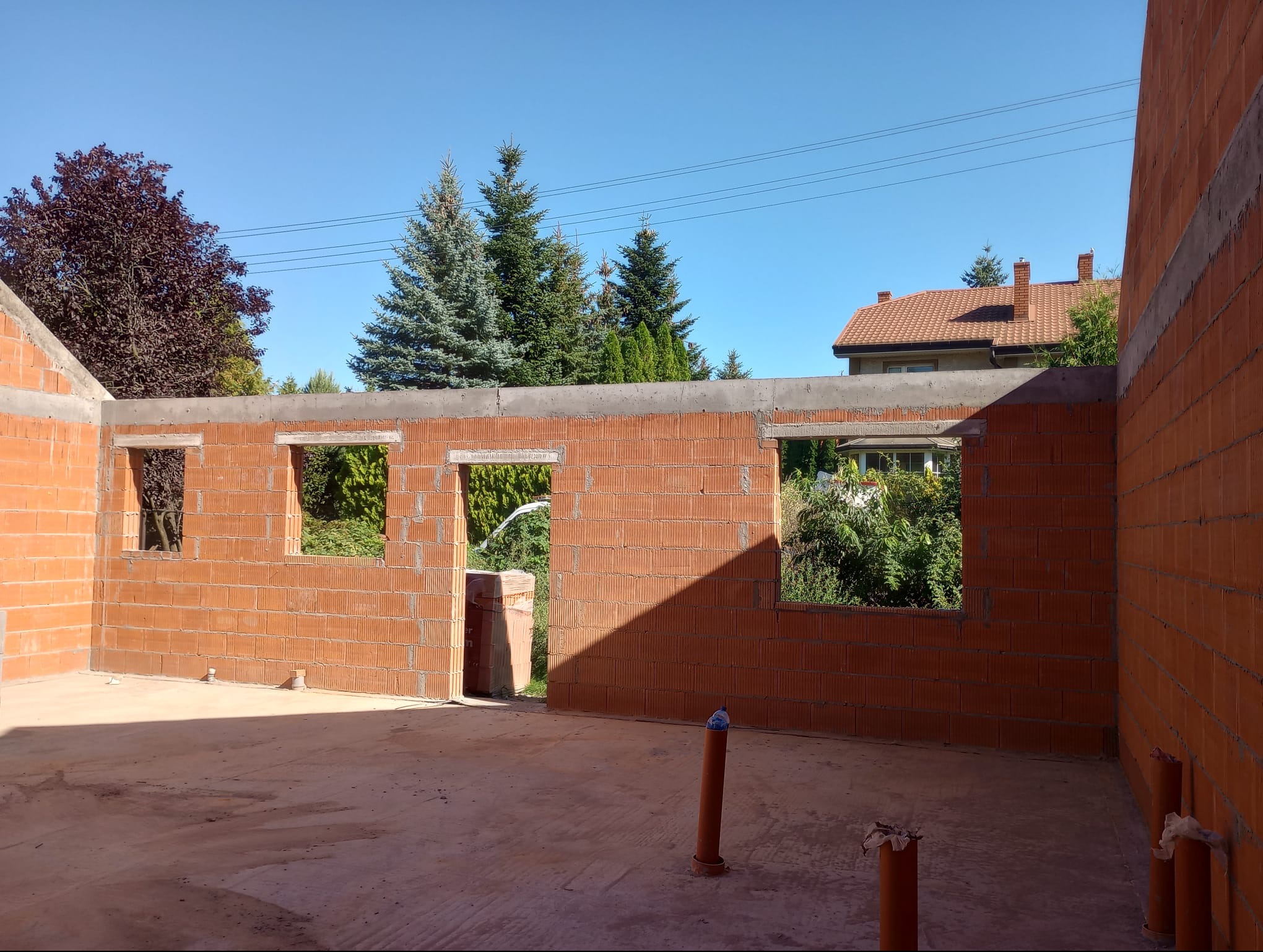 Wnętrze surowego domu z czerwonej cegły z otworami okiennymi i widokiem na zieleń, w tle błękitne niebo i dom z czerwoną dachówką. Słońce oświetla fragment ściany.