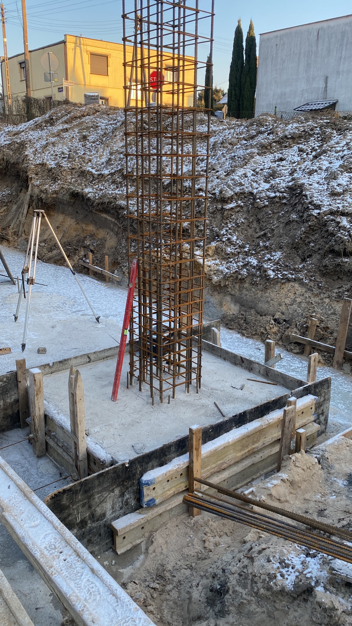 Zbrojenie kolumny na fundamencie w zasypanym śniegiem wykopie pod budowę. Widoczny niwelator i poziomica. W tle budynki i drzewa.