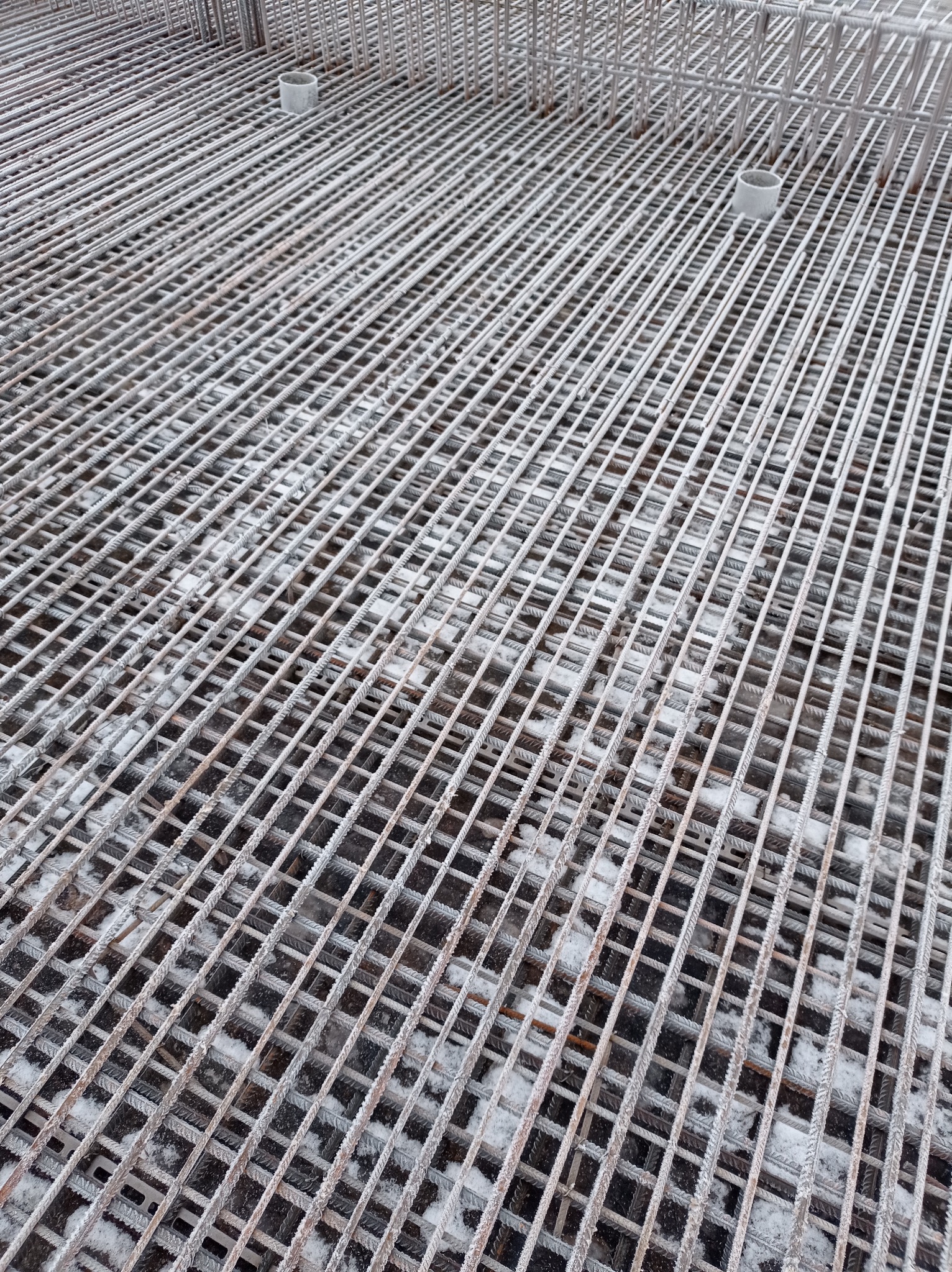 Zbrojenie fundamentów domu, widok z góry. Stalowe pręty tworzą siatkę, częściowo pokrytą szronem. Widoczne fragmenty rur instalacyjnych.