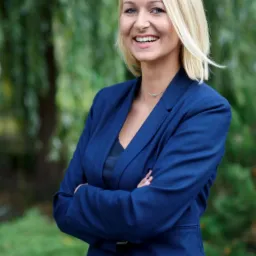 Portret uśmiechniętej blondynki w niebieskim żakiecie, skrzyżowane ręce, tło z zieleni drzew