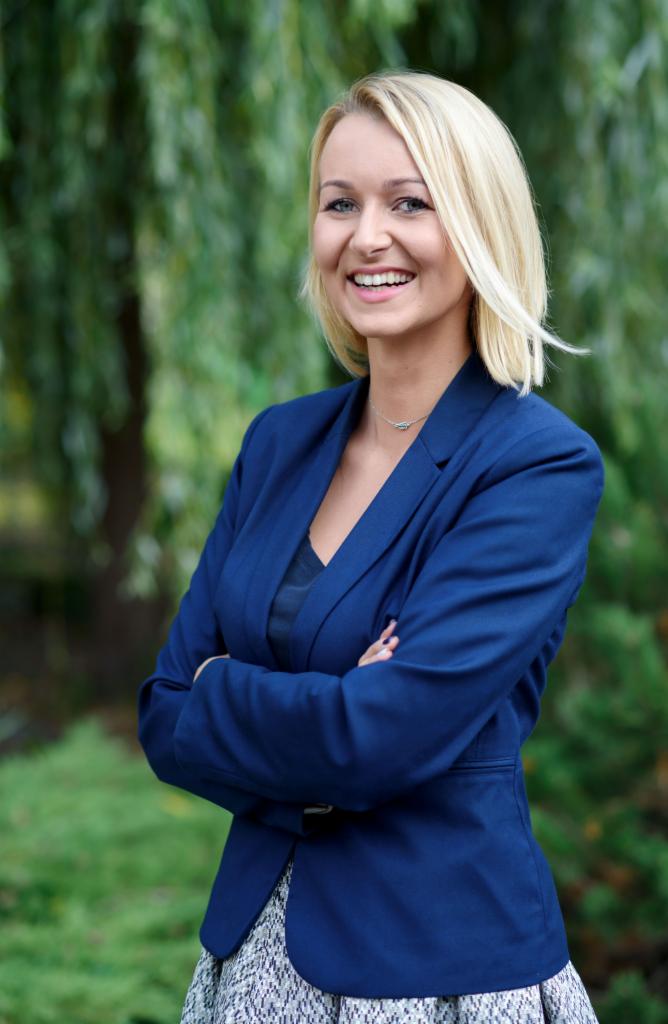 Portret uśmiechniętej blondynki w niebieskim żakiecie, skrzyżowane ręce, tło z zieleni drzew