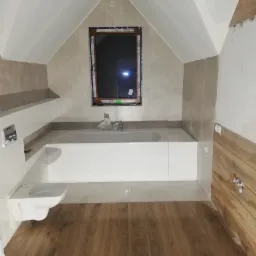 Łazienka na poddaszu w trakcie wykańczania, widoczna wanna obudowana płytkami, podwieszana toaleta, drewnopodobne panele na podłodze i okno w skosie dachu.