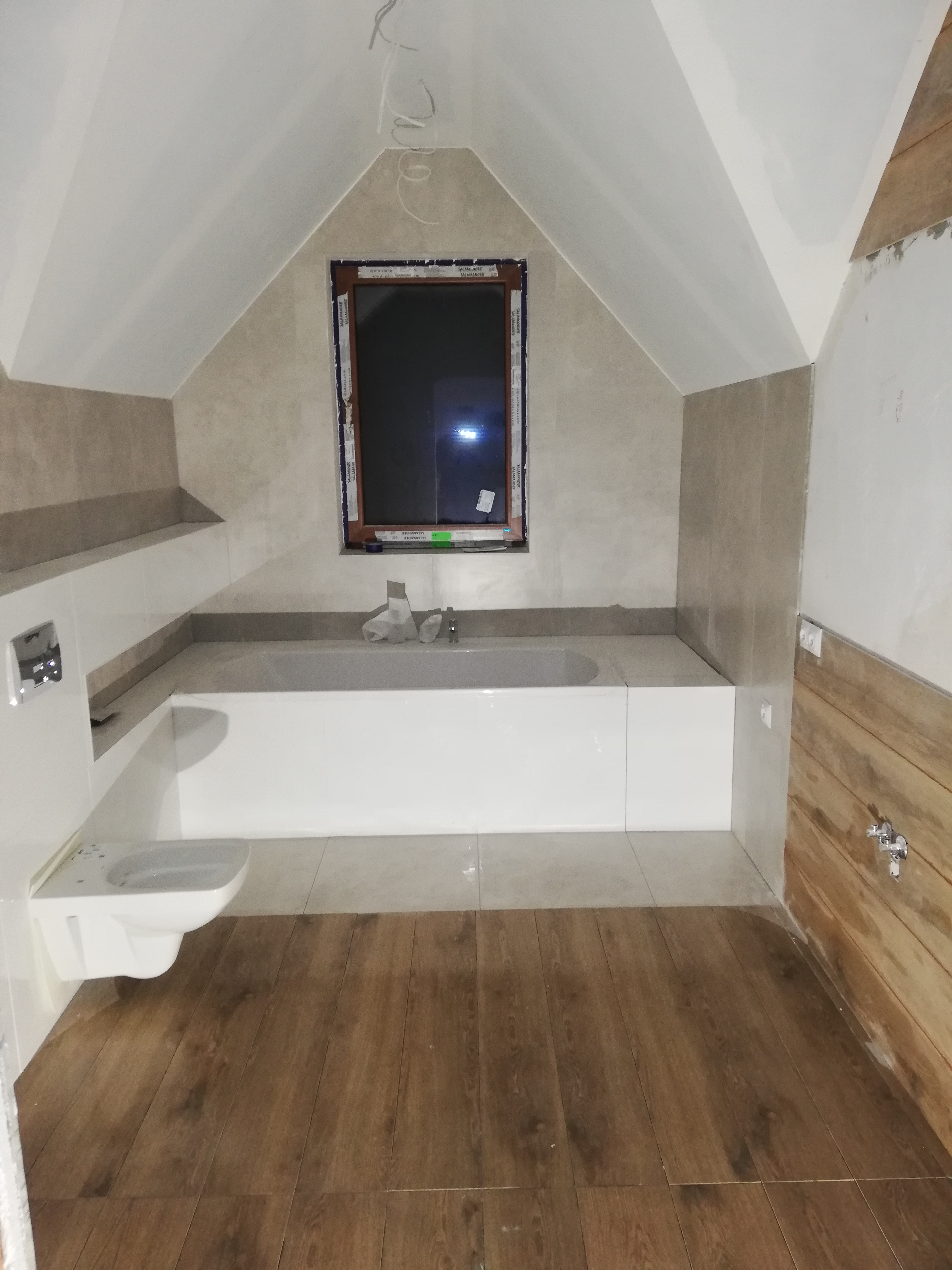 Łazienka na poddaszu w trakcie wykańczania, widoczna wanna obudowana płytkami, podwieszana toaleta, drewnopodobne panele na podłodze i okno w skosie dachu.