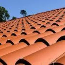 Rząd ułożonych dachówek ceramicznych w odcieniach terakoty, widok z dołu pod kątem, zieleń drzew i błękit nieba w tle.