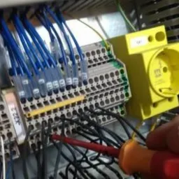 Prace elektryczne w rozdzielni: widoczne kable w kolorze niebieskim i czarnym, listwy zaciskowe, żółte gniazdo, dłoń elektryka z wkrętakiem z czerwoną rękojeścią podczas podłączania przewodów.