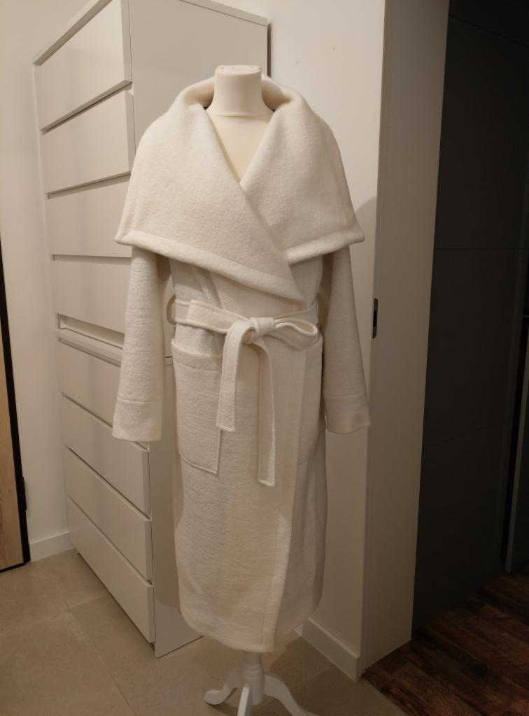 Długi, kremowy szlafrok z szerokim kołnierzem i wiązaniem w pasie, zaprezentowany na manekinie na tle białej komody i fragmentu drewnianej podłogi.